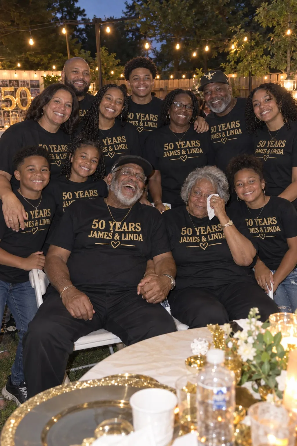 An older couple holding hands surrounded by their family in black shirts with gold “50 Years, James & Linda” letters