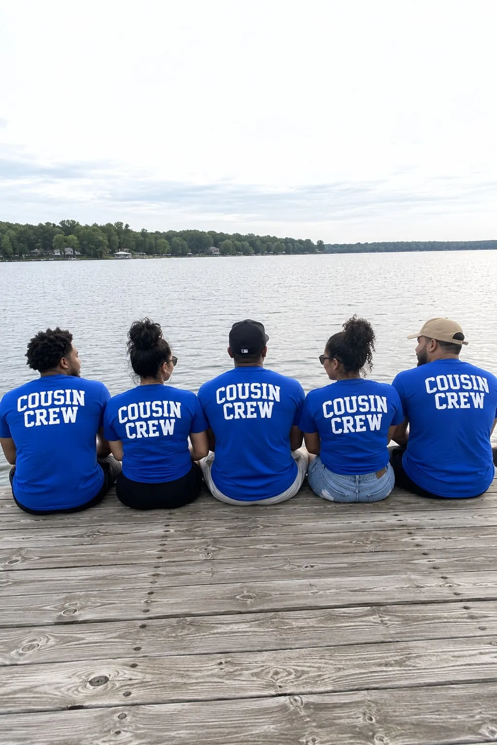 Five adult cousins sitting on a lake dock seen from behind, wearing blue shirts with “Cousin Crew” on the back