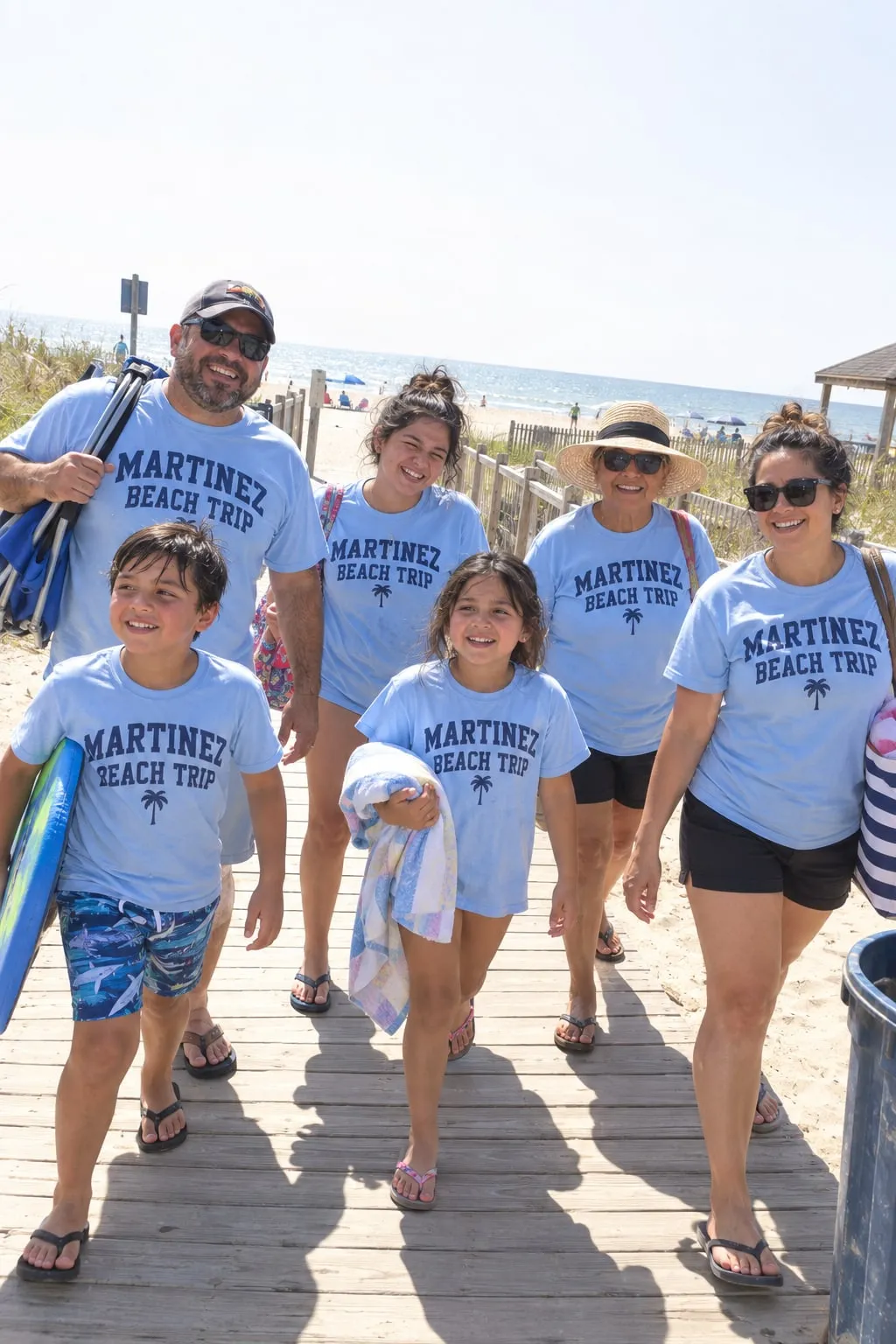 A family walking on a beach boardwalk in light blue “Martinez Beach Trip” shirts with a palm tree