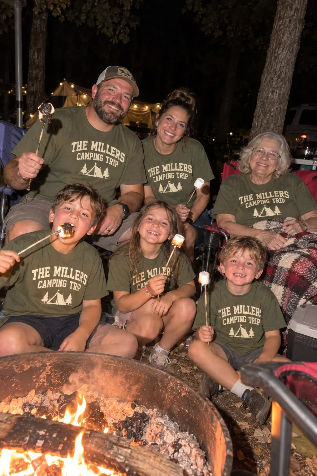 A family roasting marshmallows around a campfire at night in olive “The Millers Camping Trip” shirts