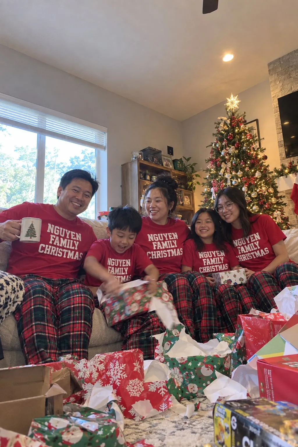 A family of five on a couch opening presents in red “Nguyen Family Christmas” shirts and plaid pajama pants