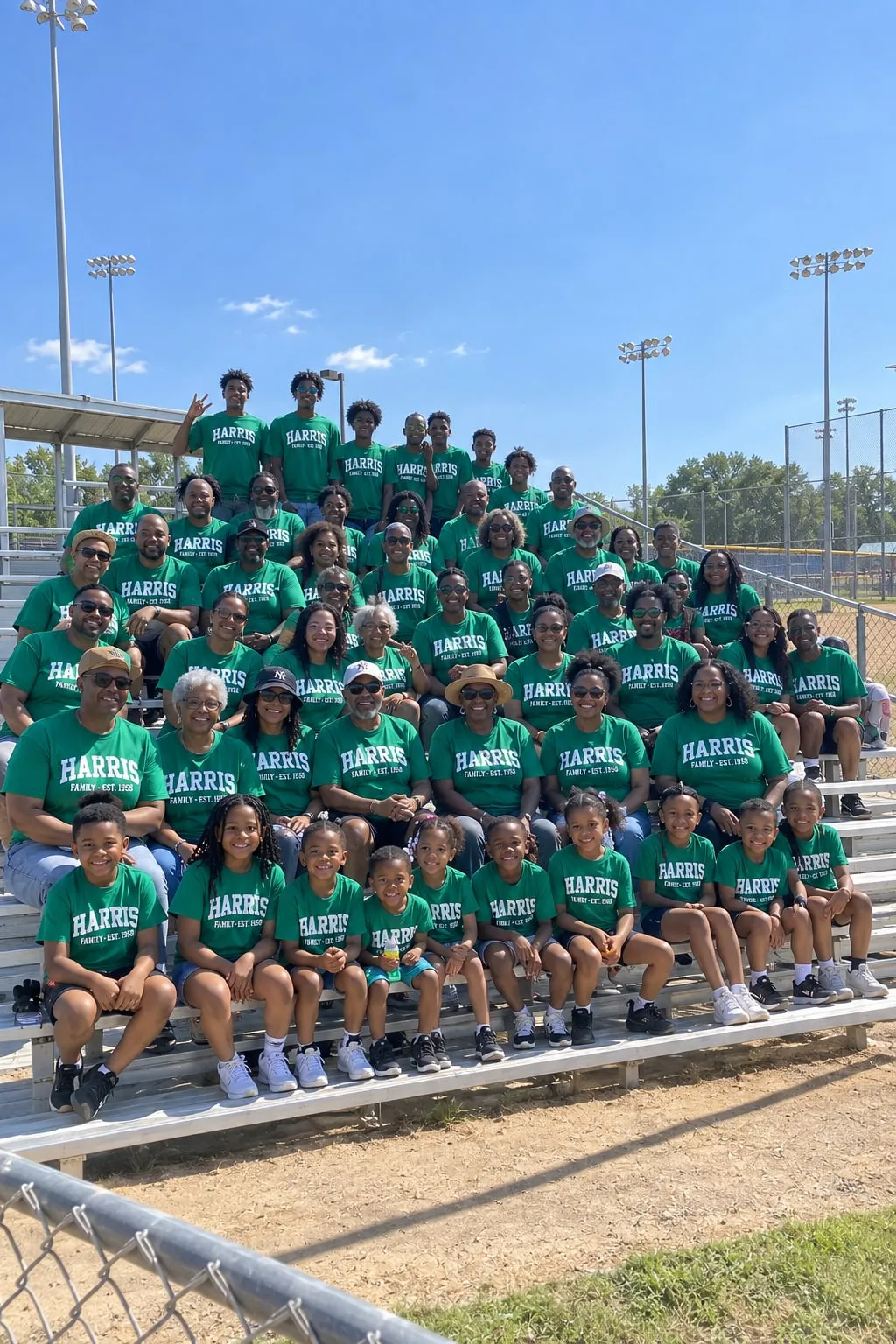 About sixty relatives on ballfield bleachers in matching green shirts reading “Harris Family, Est. 1958”