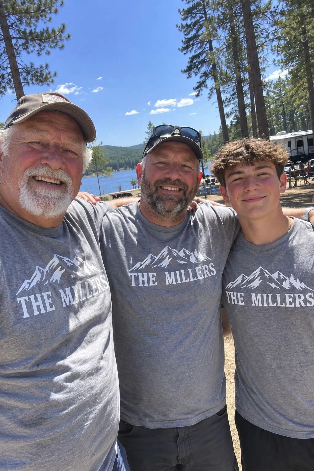 A grandfather, father and teenage son by a lake in matching grey shirts with a mountain outline and “The Millers”